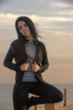 Kadın, gün batımında arka planda okyanus olan bir rıhtımda güven içinde poz veriyor. Uzun saçları rüzgarda savruluyor. Günlük atletik giysiler giyiyor, huzurlu atmosferi kucaklıyor.