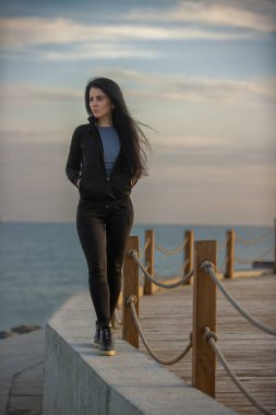 Kadın sahildeki bir kaldırımda durup güneş batarken okyanusu seyrediyor. Durgun su ve gökyüzünde sıcak bir parıltı yayıyor.