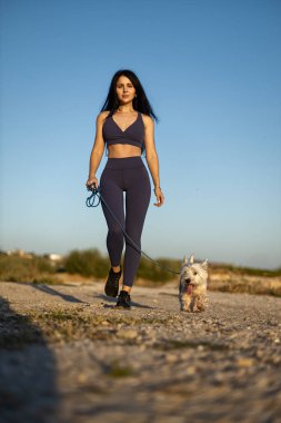 Esmer bir kadın, beyaz köpeğiyle güneşli bir sahilde geziniyor. Sıcak havanın ve okyanus esintisinin tadını çıkarırken cilt bakımına dalıyor.