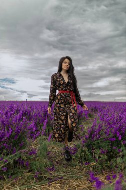 Güzel esmer yürüyüşler çiçek desenli bir elbise giymiş çiçek desenli desenli desenlerle dolu bir tarlada zarif bir şekilde yürür gökyüzü karamsar bir zemin oluşturur.