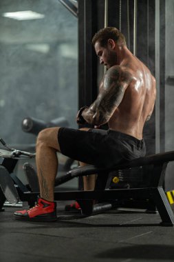 Spor salonundaki bir bankta otururken kaslı bir adam beline blok çekme hareketi yapıyor. Bu dinamik sahne güç eğitimi sırasında odak ve kararlılığı yakalar.