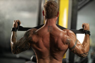 Sporcu, bir spor salonunda öne doğru çekilme hareketi yaparken gücünü gösteriyor. Kaslar gözle görülür bir şekilde bağlı ve akşam saatlerinde çalışma yoğunlaştıkça gerginlik de azalıyor..