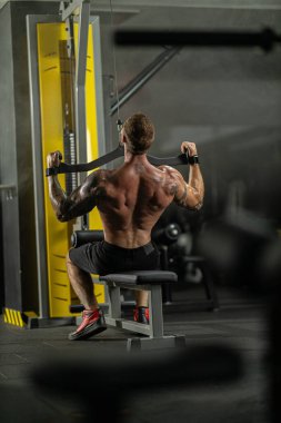 Sporcu, spor salonundaki bir bankta otururken formda olduğunu gösteren bir ön çekme egzersizi yapar. Spor salonu ayarları çeşitli egzersiz makineleriyle donatılmıştır..