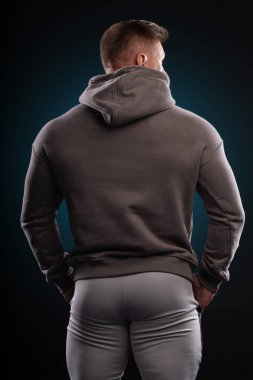 athletic man wearing a gray hoodie displays a strong physique while preparing for an evening workout. The scene is set in a studio with a dark background that highlights his silhouette.