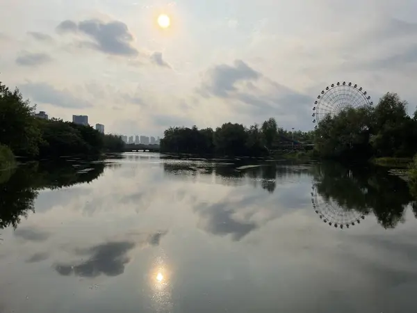 Yanghu Wetland Park, a famous tourist site in Changsha, China. Trees, sunset, ferris wheel, clouds reflected in calm lake.