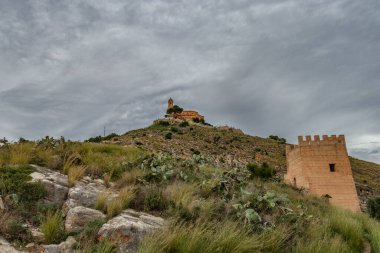 Bir yürüyüş yolunun dibindeki Cullera kalesi, etrafında kaktüsler ve kuleler olan kayalık yeşil tepe.