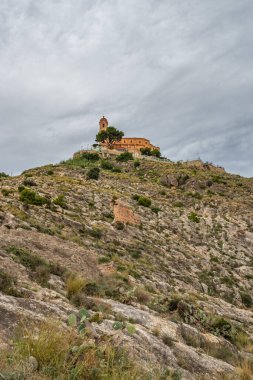 Bir yürüyüş yolunun dibindeki Cullera kalesi, etrafında kaktüsler ve kuleler olan kayalık yeşil tepe.