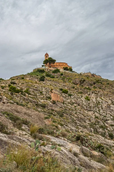 Bir yürüyüş yolunun dibindeki Cullera kalesi, etrafında kaktüsler ve kuleler olan kayalık yeşil tepe.