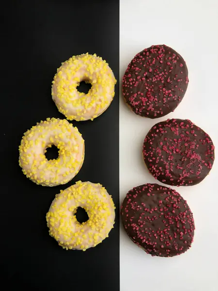 Siyah zemin üzerinde, donut, sarı ve pembe serpiştirilmiş beyaz çikolatalı çörekler; çikolata kaplı, beyaz zemin üzerinde koyu kırmızı çörekler