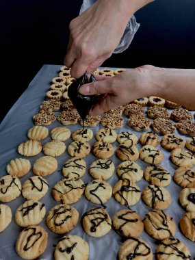 Bir insanın elleri çikolata serpmek için krema tüpü kullanıyor. Sıraların üstüne ve sıra sıra dizilmiş küçük kurabiyelerin sütunlarına.
