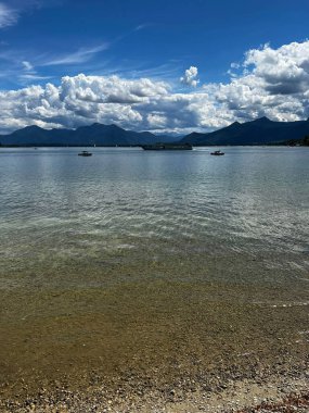 Gölün bir fotoğrafı, berrak mavi gökyüzü ve beyaz bulutlar, uzaktan belli belirsiz dağlar.