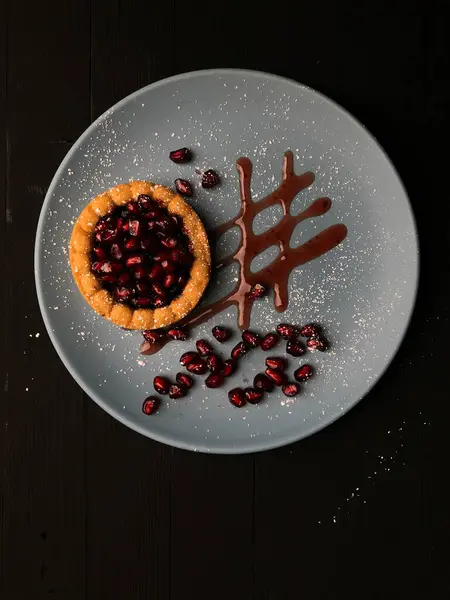 Yemek fotoğrafçılığı, nar tartlı gri bir tabak ve çikolatalı çiseleme, üst manzara, koyu arkaplan.