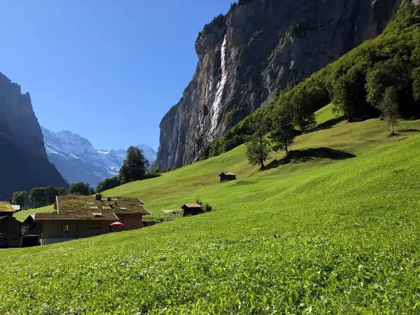 Lauterbrunnen Vadisi, İsviçre, arka planda bir şelale, yeşil çimenlik bir tarla, ve üzerinde küçük evler, açık mavi bir gökyüzü, güzel bir doğal manzara