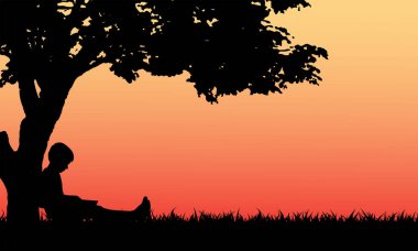 Küçük çocuk parkta ağacın altında kitap okuyor.