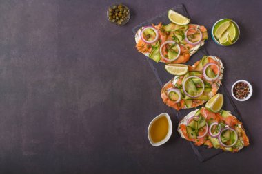Açık sandviçler, tuzlu somon, krem peynir, soğan, kapari, salatalık ve mikroyeşillikler, koyu gri arka planda. Deniz ürünleri. Sağlıklı yiyecekler. Kapatın. Üst Manzara. Boşluğu kopyala
