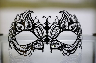 Maskeyi andıran siyah beyaz bir başlık cam bir masanın üzerinde duruyor. Tasarım, simetri ve süs unsurlarını sanatsal bir şekilde birleştiriyor.