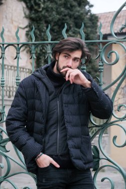 Pürüzsüz siyah deri ceketli bir adam canlı yeşil bir kapıya yaslanıyor, resmi kıyafetine serin bir güven yayıyor.