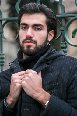 Pürüzsüz siyah deri ceketli bir adam canlı yeşil bir kapıya yaslanıyor, resmi kıyafetine serin bir güven yayıyor.