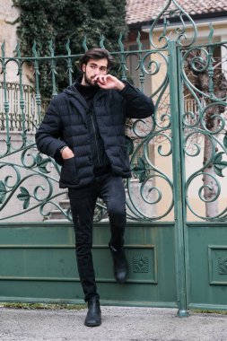 Pürüzsüz siyah deri ceketli bir adam canlı yeşil bir kapıya yaslanıyor, resmi kıyafetine serin bir güven yayıyor.