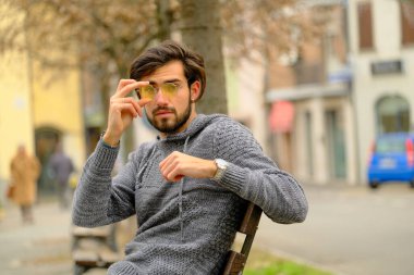 Sakallı, şapkalı ve gözlüklü bir adam kaldırımda elinde cep telefonuyla yürüyor. Kolları bir binanın önünden geçerken ağaca sürtünüyor.