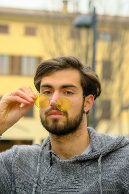 Sakallı, şapkalı ve gözlüklü bir adam kaldırımda elinde cep telefonuyla yürüyor. Kolları bir binanın önünden geçerken ağaca sürtünüyor.