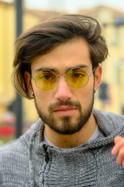 Sakallı, şapkalı ve gözlüklü bir adam kaldırımda elinde cep telefonuyla yürüyor. Kolları bir binanın önünden geçerken ağaca sürtünüyor.