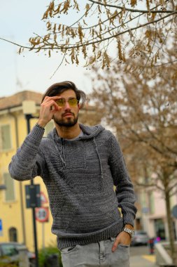 Sakallı, şapkalı ve gözlüklü bir adam kaldırımda elinde cep telefonuyla yürüyor. Kolları bir binanın önünden geçerken ağaca sürtünüyor.