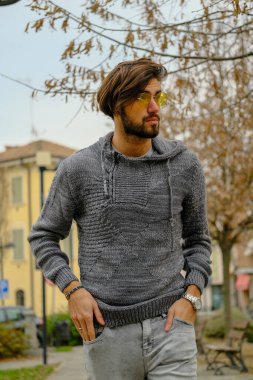 Sakallı, şapkalı ve gözlüklü bir adam kaldırımda elinde cep telefonuyla yürüyor. Kolları bir binanın önünden geçerken ağaca sürtünüyor.