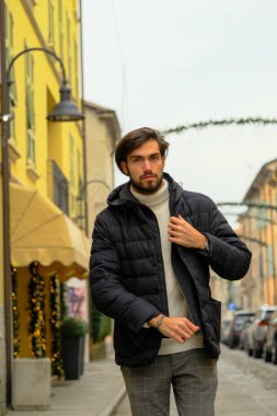 Siyah ceketli bir adam şehir caddesinde asfalt yol yüzeyi ve kaldırımı süsleyen saksılarla bina cephelerinde geziniyor.