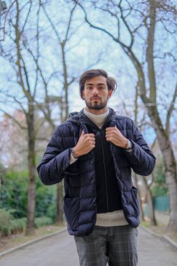 Siyah ceketli bir adam şehir caddesinde asfalt yol yüzeyi ve kaldırımı süsleyen saksılarla bina cephelerinde geziniyor.