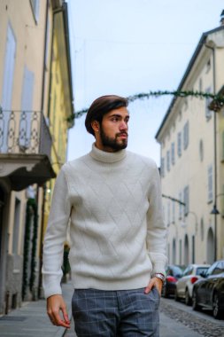 Beyaz kazak ve şapka giyen sakallı bir adam caddede yürüyor, ayaklarının altında asfalt var ve binalar onun önünde yükseliyor.