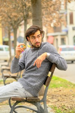 Bir adam parkta bir bankta oturuyor, bir ağacın yanında, cep telefonuyla konuşuyor. Arka planda, patlak lastikli ve pencereli park edilmiş bir araba var.