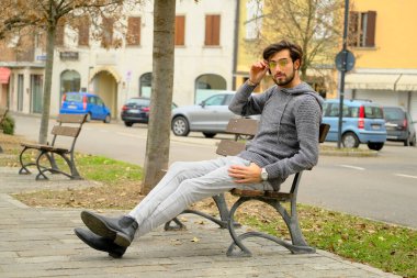 Bir adam parkta bir bankta oturuyor, bir ağacın yanında, cep telefonuyla konuşuyor. Arka planda, patlak lastikli ve pencereli park edilmiş bir araba var.