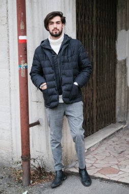 Sakallı bir adam gri bir gömlek giyiyor, kolları kıvrılmış, deri ceket ve kot pantolon giyiyor. Beton bir duvarın önünde duruyor, flaşlı fotoğraflarla sakalını ve yakasını vurguluyor.