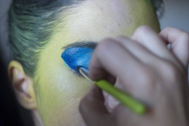 Bir insan, hassas bir fırçayla bir kadının göz kapağına mavi göz farı sürüyor, gözlerini büyütüyor ve renk katıyor.