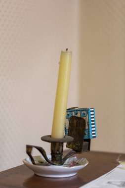 Silindir şeklinde sarı bir mum, bir masanın üzerindeki mum tutacağının içinde duruyor. Ilık bir parıltı yayıyor. Kurulum, natürmort ve iç tasarım sanatını çağrıştırıyor.