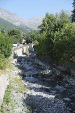 Arkaplanda görkemli dağların olduğu canlı yeşil bir ormanın içinden geçen bir nehir, sakin ve pitoresk bir doğal manzara yaratıyor.