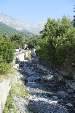 Arkaplanda görkemli dağların olduğu canlı yeşil bir ormanın içinden geçen bir nehir, sakin ve pitoresk bir doğal manzara yaratıyor.