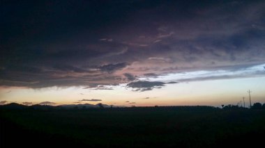 Sahne, gün batımında karanlık bulutlu bir gökyüzünün altında atmosferik ve meteorolojik fenomenlerle birlikte alacakaranlıkta doğal manzaranın güzelliğini yansıtıyor.