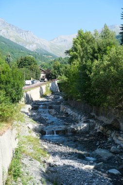 Arkaplanda görkemli dağların olduğu canlı yeşil bir ormanın içinden geçen bir nehir, sakin ve pitoresk bir doğal manzara yaratıyor.