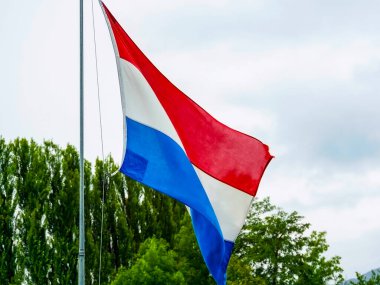 Güzel bir açık hava sahnesinde ulusal gururu sembolize eden, bulutlu bir gökyüzünde renkli Hollanda bayrağı dalgalanır.