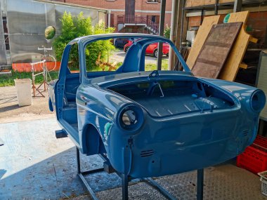 Canlı bir mavi araba, iş için gerekli araç ve ekipmanlarla çevrili geniş bir garajın içinde boyanmakta.