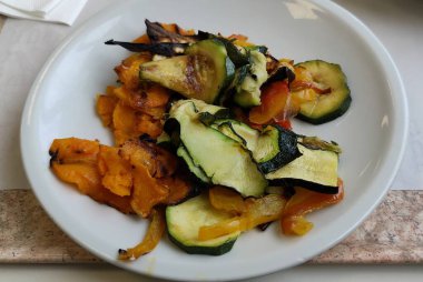 Renkli bir çeşit ızgara sebzeyle sanatsal olarak düzenlenmiş el değmemiş beyaz bir tabak. Yemek masasına güzelce yerleştirilmiş.