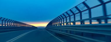 A car is steadily driving down an expansive highway while the sun sets beautifully in the background, painting the sky with vibrant colors clipart