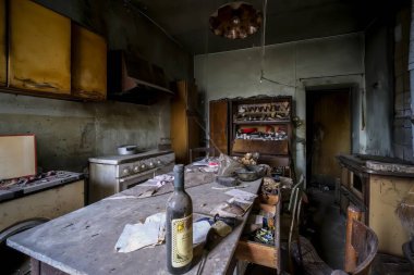 Terk edilmiş ve harap olmuş bir mutfaktaki masanın üzerine tünemiş yalnız bir şarap şişesi nostalji ve gizemi çağrıştırıyor.