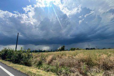 Uçsuz bucaksız yeşil bir arazide dolambaçlı bir yol ilerlerken fırtınalı bir gökyüzü arka planda kaygı verici bir şekilde beliriyor ve dramatik bir sahne yaratıyor.