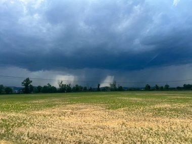 Uçsuz bucaksız yeşil bir arazide dolambaçlı bir yol ilerlerken fırtınalı bir gökyüzü arka planda kaygı verici bir şekilde beliriyor ve dramatik bir sahne yaratıyor.