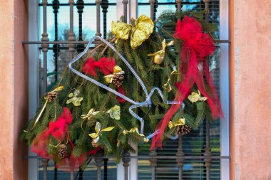 A festive Christmas wreath hangs on a window, decorated with a blue star, gold ornaments, and red ribbons. clipart