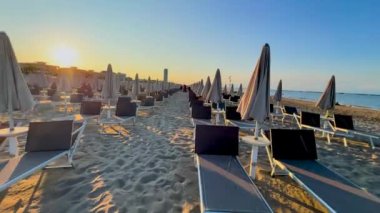 Altın saat sıcak ışıkta Cesenatico plajında yıkanır, kumsalda ve plaj sandalyelerinde uzun gölgeler bırakır. Adriyatik kıyısında sakin bir manzara.