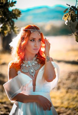 Çekici kızıl saçlı bir kadın zarif bir şekilde beyaz bir gelinlik ve görünüşünü güzelleştiren çarpıcı bir kolye giyiyor.